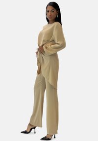 Beige satin sæt med en langærmet top med en sideløkke og brede bukser, parret med sorte spidse hæle.