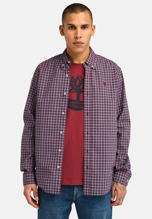 Camisa a cuadros roja y azul con botones y cuello, llevada sobre una camiseta gráfica roja con un diseño de árbol. Corte casual, tejido ligero.