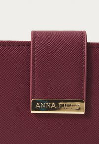 Geantă din material sintetic texturat de culoare burgundă, cu un plăcuță de metal aurie cu logo-ul "ANNA FIELD" pe clapeta superioară. Design elegant și funcțional.