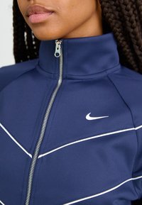 Mornarški zip-up jopič z visokim ovratnikom, belimi akcentnimi črtami in majhnim Nike logotipom na prsih. Gladka tekstura, športni dizajn.