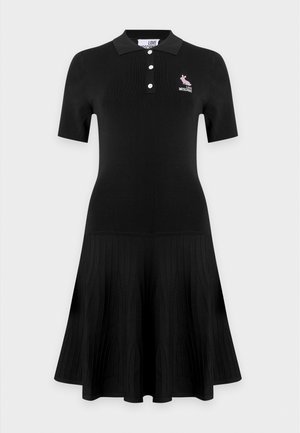Jeune femme portant une robe noire à manches courtes avec un logo, des bottes noires montantes avec des boutons, tenant un petit sac rouge.