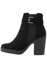 Botines de ante negros con un tacón ancho y bloque, paneles laterales elásticos y un acento de hebilla plateada en la correa del tobillo. Suela de goma texturizada.
