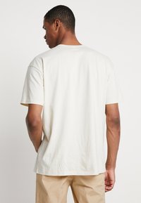 T-shirt in cotone beige chiaro con maniche corte, scollo a girocollo e vestibilità rilassata. Il tessuto ha una texture liscia e dettagli minimi.