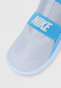 Detailní záběr na světle šedou a modrou tenisku Nike s prodyšným síťovaným svrškem a modrým popruhem se bílým logem Nike.