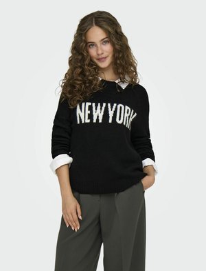 Pull en maille noire avec un motif "NEW YORK" en blanc, associé à une chemise blanche à col. Le mannequin porte un pantalon gris ajusté, coupe décontractée.