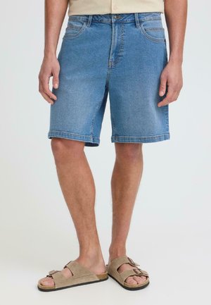 Mężczyzna noszący niebieskie szorty jeansowe o długości do połowy uda oraz beżowe sandały na otwartym palcu z dwoma paskami na klamry, stojący na tle jednolitym.