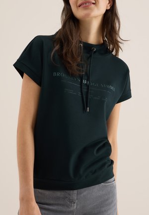 Haut à manches courtes vert foncé avec capuche et cordon de serrage, présentant un design graphique en texte sur le devant et une coupe décontractée. Texture de tissu lisse.