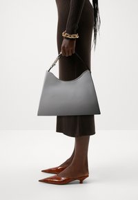 Szara skórzana torebka w kształcie półksiężyca, o gładkiej fakturze i krótkim czarnym uchwycie. Ma złoty zatrzask. Nosi się ją z prążkowaną brązową sukienką i brązowymi szpilkami.