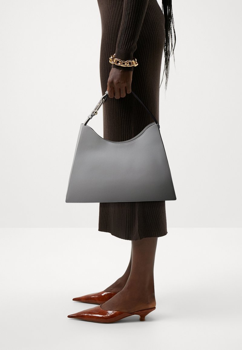 Szara skórzana torebka w kształcie półksiężyca, o gładkiej fakturze i krótkim czarnym uchwycie. Ma złoty zatrzask. Nosi się ją z prążkowaną brązową sukienką i brązowymi szpilkami.