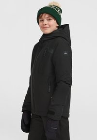 Zwarte geïsoleerde jas met capuchon, ritszakken en logo. G gedragen met zwarte wanten en een groene gebreide beanie met een pompon bovenop.