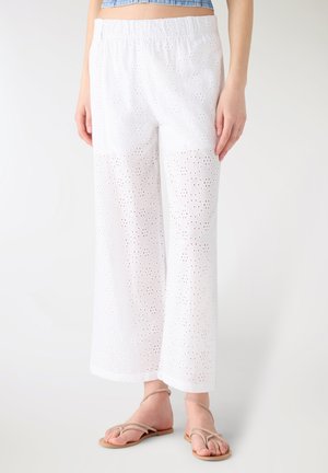 Pantaloni bianchi a gamba larga con ricamo a occhielli, vita elasticizzata, indossati con sandali beige con cinturini e un top corto blu, in piedi su un pavimento bianco.