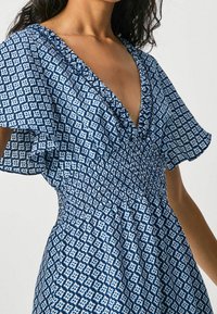 Modrá šaty s vzorem, které mají hluboký výstřih do V, volánové rukávy a nařasený pas. Tkanina má geometrický design v kontrastních odstínech.