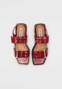 Red patent leather sandals featuring a square toe, wide straps with grommets, and a silver buckle accent. Flat sole with a textured finish.