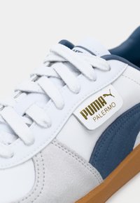 Bijela tenisica s svijetlo sivim nubuk detaljima, plavim bočnim panelom i zlatnim Puma logom. Ima bijele vezice i gumeni potplat boje drveta.