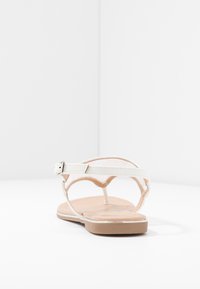 Valkoinen sandaali, jossa on litteä pohja, avoin takaosa ja ohuet hihnat. Varustettu solkikiinnityksellä ja pehmustetulla ruskealla sisäpohjalla. Sileä pinta.