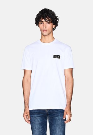 Maglietta bianca in cotone con scollo rotondo, caratterizzata da un piccolo logo rettangolare nero con scritte dorate sulla tasca sinistra del petto.