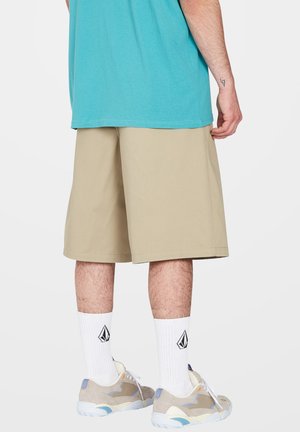 Beige Shorts mit lockerer Passform, kombiniert mit grauen und blauen Sneakers, weißen Ripp-Socken und einem türkisfarbenen T-Shirt, von hinten betrachtet.