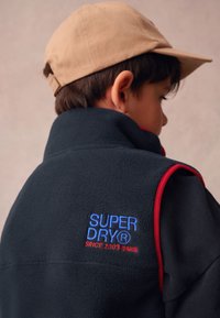 Juodas vilnos liemenė su raudonomis detalėmis; nugaroje siuvinėtas "SUPERDRY" logotipas mėlyna ir raudona spalvomis; derinamas su smėlio spalvos kepure.