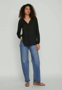 Woman with curly hair wearing a black long-sleeve blouse, blue jeans, and beige flats, standing with one hand in pocket against plain background.