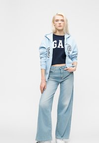 Jeune femme aux cheveux blonds portant un sweat à capuche GAP bleu clair, un t-shirt court bleu marine, un jean large bleu clair et des baskets blanches, debout.