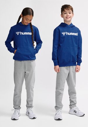 Blauwe hoodie met witte "hummel" logo, gecombineerd met lichtgrijze joggingsbroek. Modellen dragen witte sportschoenen. Glad stofoppervlak.