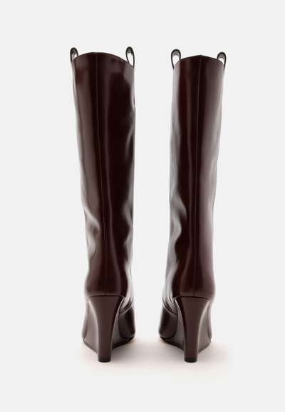 Paire de bottes en cuir marron foncé, hautes et brillantes, avec des talons compensés hauts, vues de dos sur un fond clair uni.