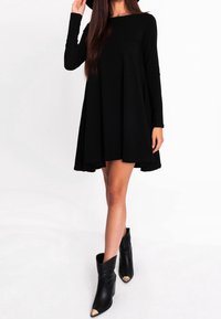 Femme portant une robe noire ample à manches longues, des bottes noires à bout pointu avec embouts dorés, et un chapeau noir, debout sur un fond blanc.