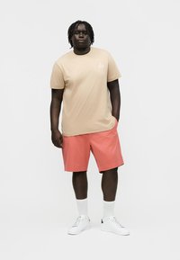 Mann med dreadlocks står iført en beige t-skjorte, korallfargede shorts, hvite crew-sokker og hvite sneakers mot en ensfarget bakgrunn.