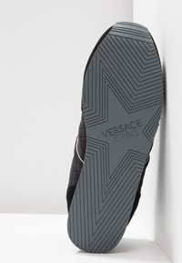 Semelle de basket noire avec un motif d'étoile géométrique et l'inscription "Versace Jeans" en relief au centre, reposant contre un coin blanc.