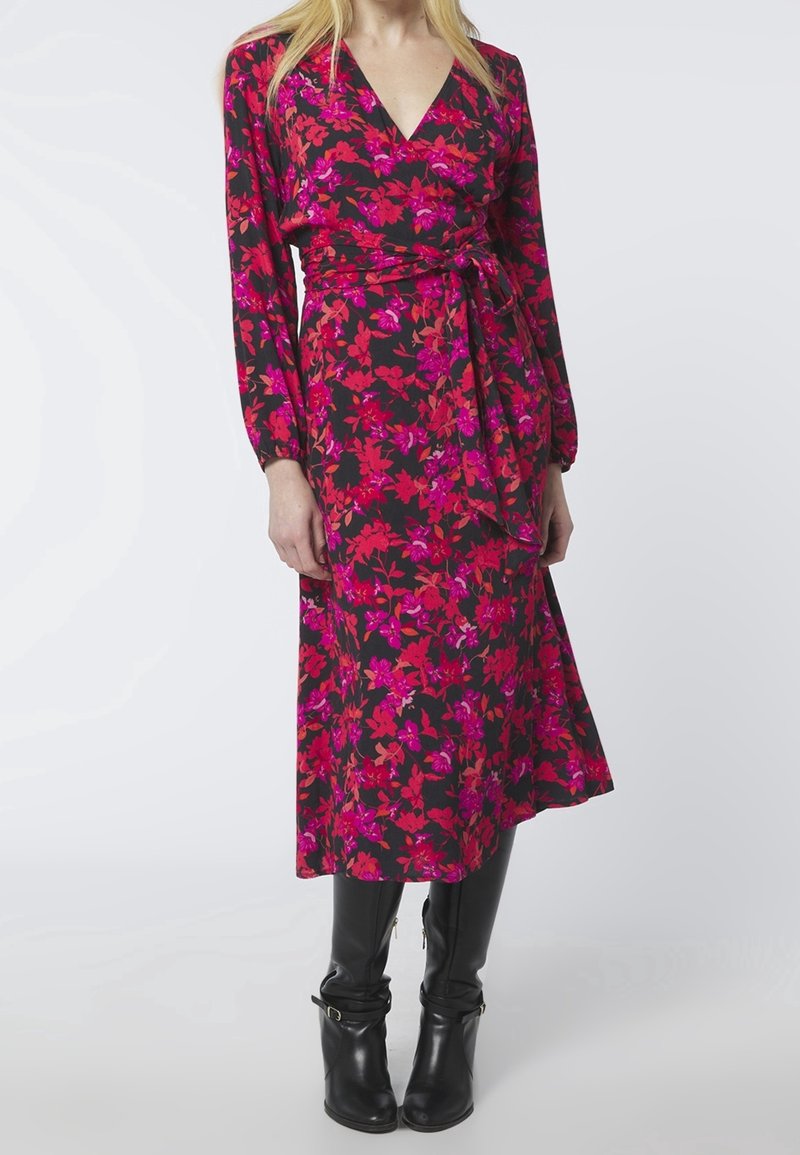 Femme portant une robe noire à manches longues avec des motifs floraux rouges et roses, une ceinture à la taille et des bottes noires montant jusqu'aux genoux.