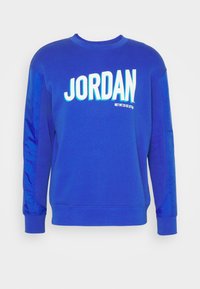 Blå sweatshirt med rund halsringning, långa ärmar och en glänsande yta på ärmarna. Har fet vit text "JORDAN" på framsidan.