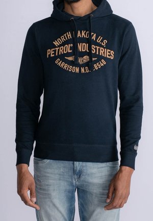 Mann trägt eine marineblaue Kapuzenjacke mit dem Text "North Dakota U.S. Petrol Industries Garrison N.D. 8540" und hellblaue Jeans, steht vor einem schlichten Hintergrund.