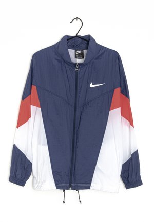 Giacca Nike con zip blu navy con pannelli a chevron rossi e bianchi, polsini elasticizzati e piccolo logo Nike Swoosh bianco sul petto.