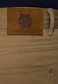 Beżowa tkanina bawełniana ze zszytymi szwami, z brązową skórzaną łatą z tłoczonym napisem "LANCERTO" oraz dekoracyjnym herbem nad tylną kieszenią.