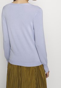 Woman wearing a light purple textured long-sleeve sweater and a mustard yellow pleated skirt, shown from the back against a plain background.