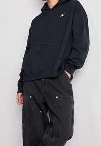 Sudadera negra con capucha y cordón, con un pequeño logotipo blanco. Combinada con pantalones cargo oscuros, que muestran detalles en color dorado.