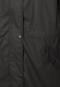 Zwarte jas close-up met voor drukknoopsluitingen en een zijflapzak met drukknoopsluiting.