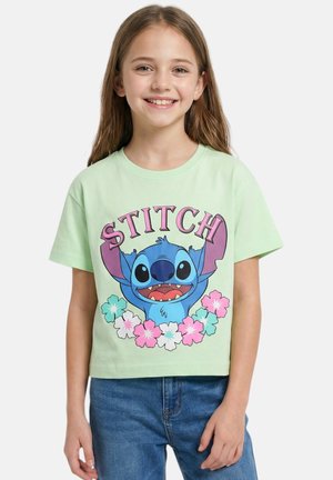Jeune fille souriante, portant un t-shirt vert clair avec le personnage de dessin animé Stitch en bleu entouré de fleurs roses et blanches, associé à un jean bleu.