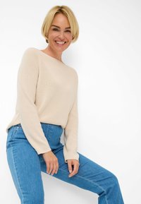 Pull en tricot beige avec un col bateau, des manches longues et une coupe décontractée, associé à un jean taille haute bleu clair. Texture lisse, design simple.