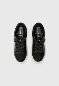 Black patent leather sneakers featuring a thick white sole, textured black laces, and a logo tag on the tongue. Smooth surface and rounded shape.