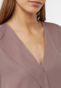 Bluzka w kolorze mauve z dekoltem w kształcie V, gładką teksturą i subtelnym przeszyciem wzdłuż krawędzi. Zbliżenie ukazujące tkaninę i kształt.