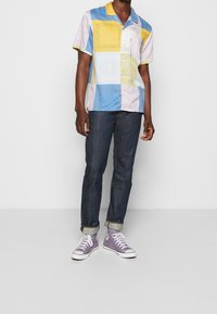 Korte mouwen shirt in een multicolor patchworkontwerp met gele, blauwe en witte panelen; gecombineerd met donkere spijkerbroek en paarse hoge sneakers.