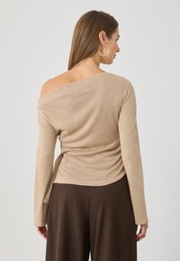 Beżowy sweter typu off-the-shoulder z długimi rękawami, o miękkiej teksturze i marszczonym szczególe z boku, w połączeniu z ciemnobrązowymi spodniami.