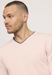 Pull à col en V en tissu rose pâle, avec un col côtelé bleu marine et un petit logo sur la poitrine, offrant une coupe décontractée.