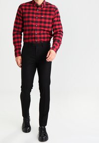 Mężczyzna w czerwono-czarną kratkę koszuli z długim rękawem, czarnych dopasowanych spodniach oraz czarnych sznurowanych butach, idzie do przodu.