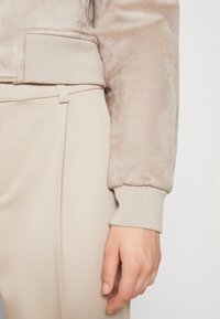 Beżowa pluszowa kurtka z prążkowanym mankietem, zestawiona z jasnobeżowymi, dopasowanymi spodniami. Tekstury są miękkie, gładkie i delikatnie kontrastujące.