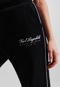 Black sweatpants made of soft fabric, featuring white piping along the side and an embroidered logo on the thigh area.