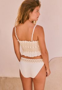 Sieviete ar viļņainiem matiem, tērpusies bēša rakstaina īsa krekla un augsta vidukļa apakšdaļa komplektā, stāv pie gaiši krāsotas sienas un raugās pa labi.