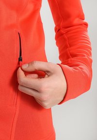 Chaqueta atlética roja con cremallera de longitud completa y bolsillo lateral. El material tiene una textura suave, forma ajustada y detalles de costura prominentes.