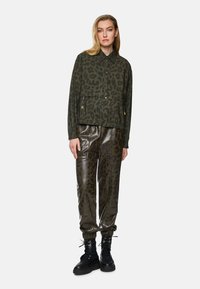 Cropped olive green jacket with black leopard print, featuring a button front and zippered pockets, paired with shiny, patterned olive pants.